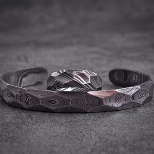 Op de afbeelding: Een set van een manchetarmband en ring van getextureerd, patroonmetaal. De open armband en ring hebben een geometrisch ontwerp. De set wordt gepresenteerd op een grijze ondergrond.
