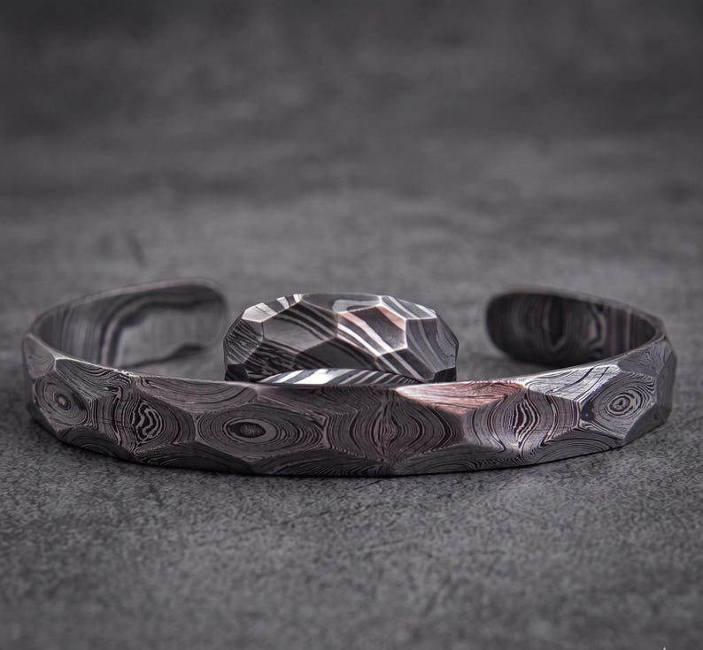 Op de afbeelding: Een set van een manchetarmband en ring van getextureerd, patroonmetaal. De open armband en ring hebben een geometrisch ontwerp. De set wordt gepresenteerd op een grijze ondergrond.