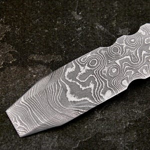 Può includere: Primo piano di una lama di coltello con un motivo a vortice unico. La lama ha un bordo strutturato e ondulato e una finitura metallica lucida. Il design intricato crea una trama visiva, suggerendo un oggetto artigianale di alta qualità.