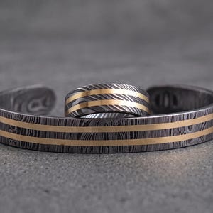 Op de afbeelding: Een bijpassende sieradenset met een manchetarmband en een ring. De armband en ring hebben een donkere, metalen basis met een patroon en goudkleurige strepen. De ring ligt op de armband.