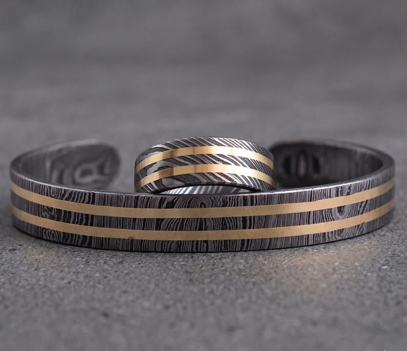 Op de afbeelding: Een bijpassende sieradenset met een manchetarmband en een ring. De armband en ring hebben een donkere, metalen basis met een patroon en goudkleurige strepen. De ring ligt op de armband.