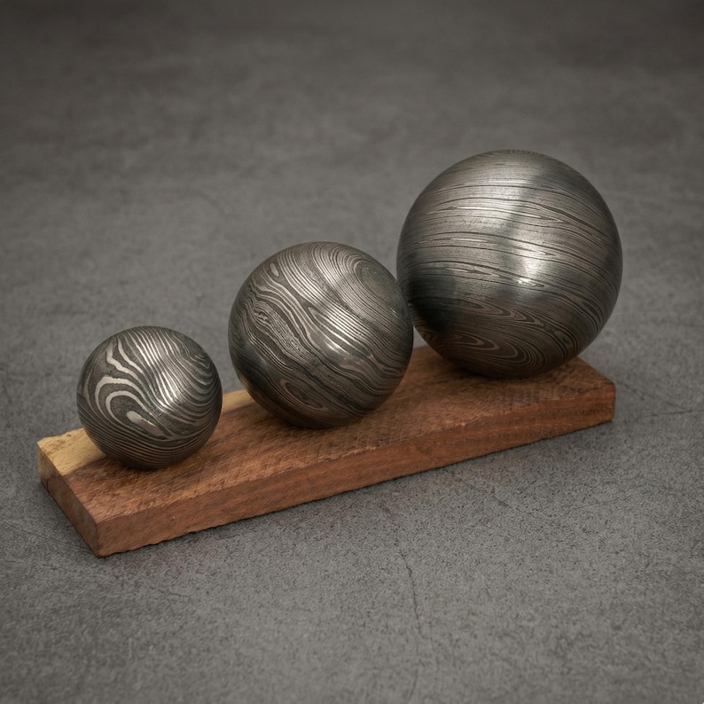 May include: Three metal spheres with a unique wood grain pattern rest on a rectangular wooden base. The spheres vary in size, with the largest on the right. The metal has a silver and dark grey color. The wooden base is a warm brown.