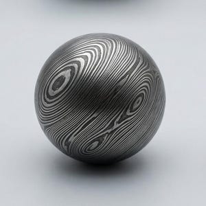 May include: A spherical metal object with a unique pattern of swirling lines. The surface appears to be made of layered metal, creating a wood-grain effect. The ball is a dark gray color and sits on a white background.