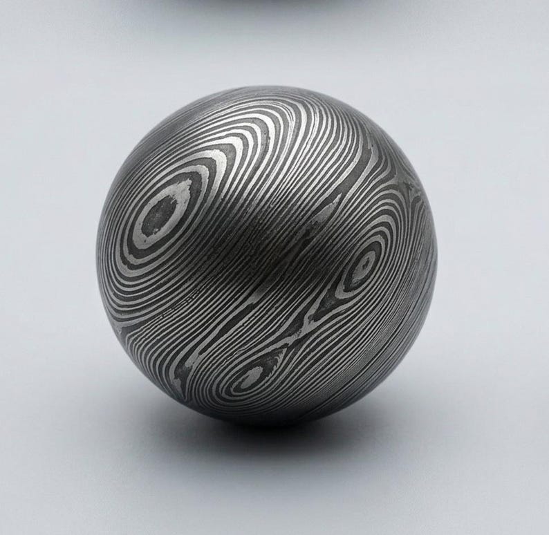 May include: A spherical metal object with a unique pattern of swirling lines. The surface appears to be made of layered metal, creating a wood-grain effect. The ball is a dark gray color and sits on a white background.