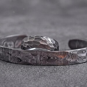 Op de afbeelding: Een zilverkleurige metalen manchetarmband en ring set. De ring heeft een facetgeslepen oppervlak, terwijl de armband en ring een uniek, wervelend patroon hebben. De sieraden worden gepresenteerd op een grijze ondergrond.
