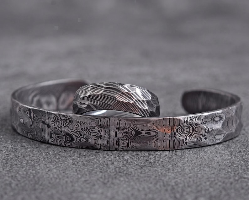 Op de afbeelding: Een zilverkleurige metalen manchetarmband en ring set. De ring heeft een facetgeslepen oppervlak, terwijl de armband en ring een uniek, wervelend patroon hebben. De sieraden worden gepresenteerd op een grijze ondergrond.