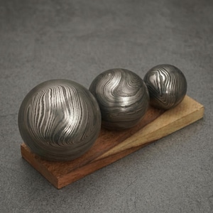 May include: Three metal spheres with a unique, swirling pattern rest on a wooden base. The spheres vary in size, with a polished, metallic finish. The wooden base has a natural wood grain and a warm, reddish-brown hue.