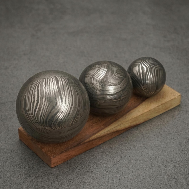 May include: Three metal spheres with a unique, swirling pattern rest on a wooden base. The spheres vary in size, with a polished, metallic finish. The wooden base has a natural wood grain and a warm, reddish-brown hue.