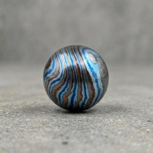 May include: A small, spherical object with a unique pattern of swirling blue, brown, and silver metallic bands. The bands create a marbled effect across the surface of the ball. The object is set against a neutral gray background.