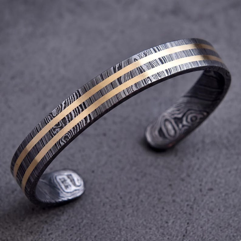 Op de afbeelding: Een manchetarmband met een patroon, donkergrijze en zilveren metalen band. Twee parallelle goudkleurige banden lopen over de bovenkant. De armband heeft een open ontwerp en wordt weergegeven op een grijze ondergrond.