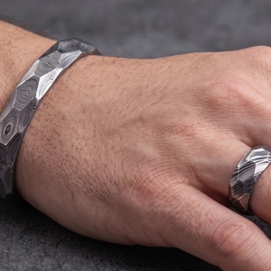 Op de afbeelding: Een metalen armband- en ringenset met patroon. De armband is een dikke band met een facetontwerp. De ring heeft een vergelijkbaar patroon en ontwerp. Het metaal heeft een donker, wervelend patroon, wat een unieke en getextureerde uitstraling cre&euml;ert.