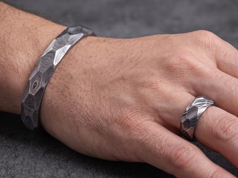 Op de afbeelding: Een metalen armband- en ringenset met patroon. De armband is een dikke band met een facetontwerp. De ring heeft een vergelijkbaar patroon en ontwerp. Het metaal heeft een donker, wervelend patroon, wat een unieke en getextureerde uitstraling cre&euml;ert.