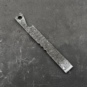 以下が含まれることがあります： 渦巻くグレーと黒のラインが特徴的なダマスカス鋼模様の平らで細長い金属製ツール。一端に円形の穴があり、側面には刻み目があり、反対側の端は真っ直ぐで平らな形状をしています。