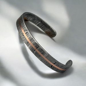 May include: A Damascus steel cuff bracelet with a copper inlay. The bracelet has a textured, patterned surface in shades of gray and black, with a central copper stripe. The open-ended design allows for easy wear.