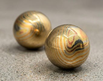 May include: Two metallic spheres with a swirling pattern of gold, bronze, and silver tones. The spheres are smooth and reflective, resting on a textured gray surface. The spheres appear to be decorative objects.