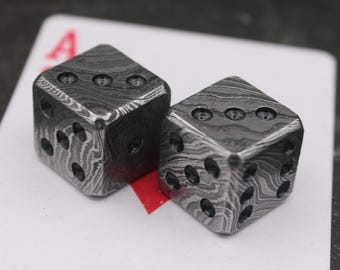 May include: Two metal dice with a unique patterned design, resting on a playing card. The dice are dark gray with black dots indicating the numbers. The playing card has a red 'A' on it.