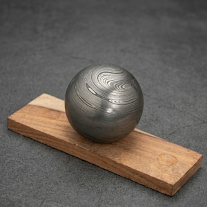 Puede incluir: Una esfera metálica con un patrón arremolinado único descansa sobre una tabla de madera rectangular. La superficie de la esfera tiene un aspecto texturizado y en capas, con tonos de gris y plata. La tabla de madera es marrón claro.