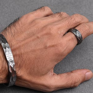 Op de afbeelding: Een getextureerde, donkere metalen manchetarmband en ring set. De armband is een brede band met een gehamerd patroon. De ring heeft een vergelijkbaar ontwerp, beide lijken gemaakt van een donker, mogelijk geoxideerd metaal. De sieraden worden op een hand getoond.