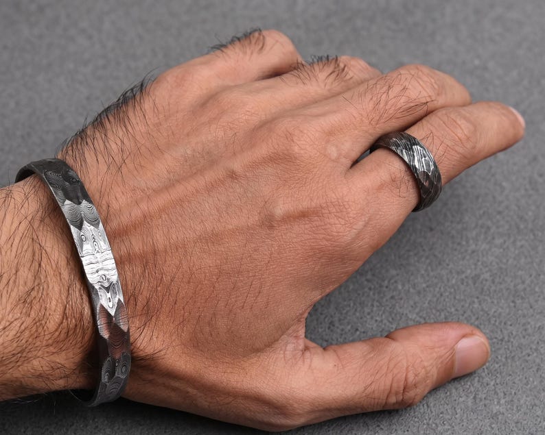 Op de afbeelding: Een getextureerde, donkere metalen manchetarmband en ring set. De armband is een brede band met een gehamerd patroon. De ring heeft een vergelijkbaar ontwerp, beide lijken gemaakt van een donker, mogelijk geoxideerd metaal. De sieraden worden op een hand getoond.