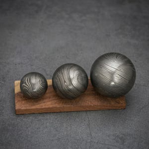 May include: Three metal spheres with a unique, swirling pattern rest on a rectangular wooden base. The spheres vary in size, with a dark, metallic finish. The wood base is a warm brown color, providing a contrast to the spheres.
