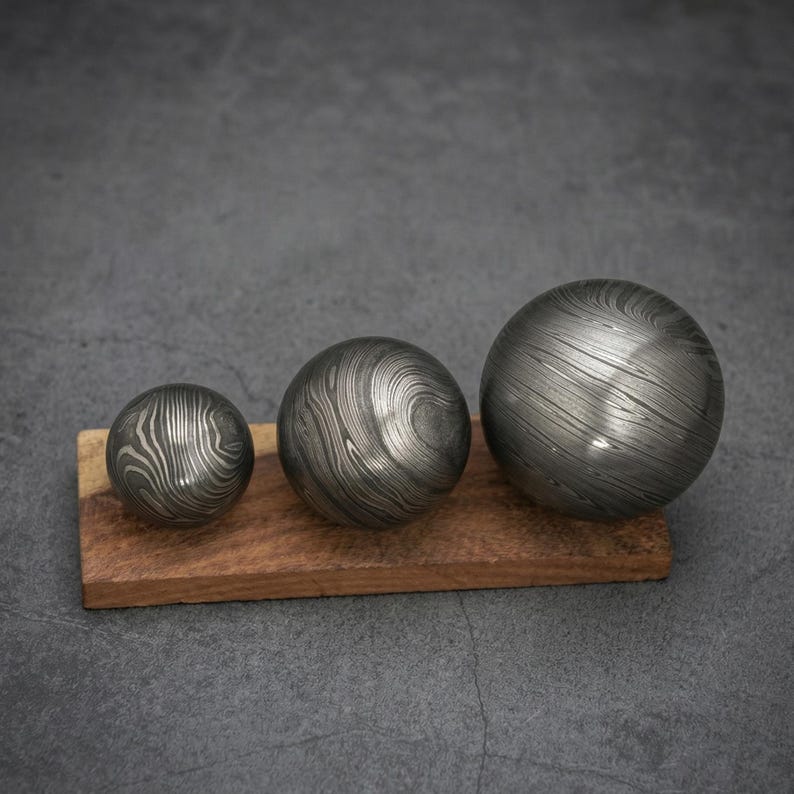 May include: Three metal spheres with a unique, swirling pattern rest on a rectangular wooden base. The spheres vary in size, with a dark, metallic finish. The wood base is a warm brown color, providing a contrast to the spheres.