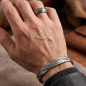 Op de afbeelding: Een getextureerde zilveren ring en bijpassende manchetarmband. De ring zit aan een vinger en de armband om een pols. De sieraden hebben een uniek patroon en een donkere, metallic afwerking. De achtergrond is wazig.