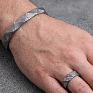 Op de afbeelding: Een zilverkleurige armband- en ringenset. De armband is een dikke, geometrische manchet. De ring heeft een bijpassend geometrisch ontwerp. Beide stukken hebben een textuur, patroon oppervlak. De sieraden worden op een hand getoond.