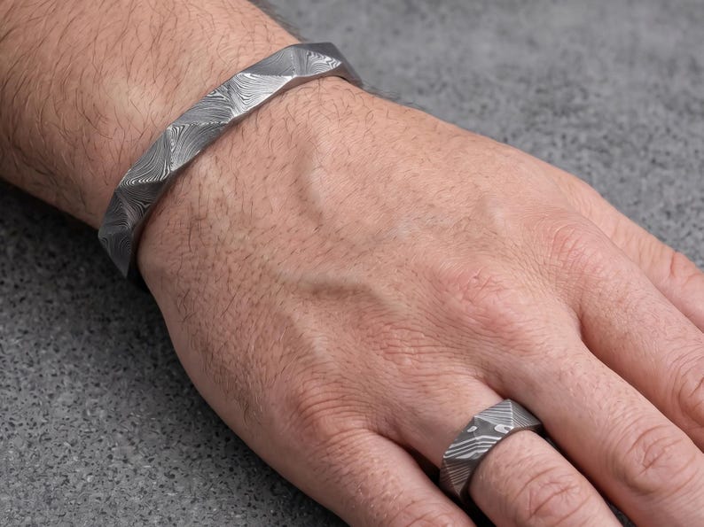 Op de afbeelding: Een zilverkleurige armband- en ringenset. De armband is een dikke, geometrische manchet. De ring heeft een bijpassend geometrisch ontwerp. Beide stukken hebben een textuur, patroon oppervlak. De sieraden worden op een hand getoond.