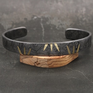 Puede incluir: Una pulsera de puño de metal gris oscuro con un diseño estampado y acentos triangulares dorados. La pulsera se exhibe sobre un pequeño soporte de madera, mostrando sus intrincados detalles y artesanía.