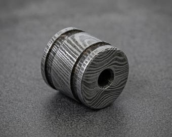 May include: A cylindrical metal bead with a Damascus steel pattern. The bead has a central hole and is made of a dark, textured metal. The design features a series of concentric rings and wavy lines.