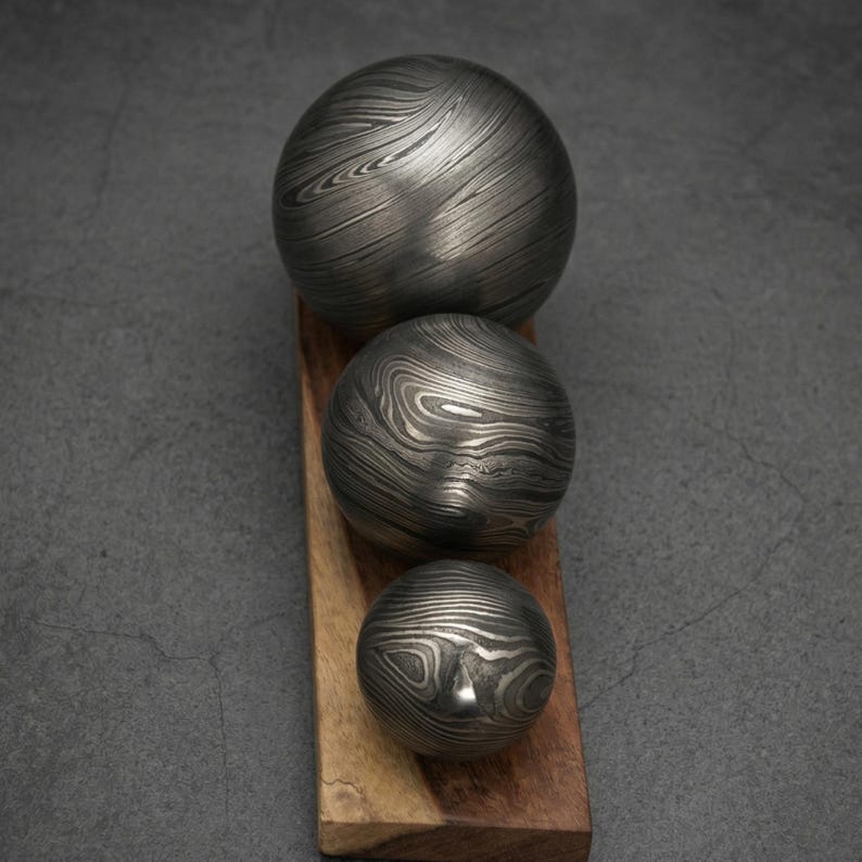 May include: Three decorative metal spheres with a unique swirling pattern, arranged on a wooden base. The spheres vary in size, with the largest at the top and the smallest at the bottom. The metal has a dark, textured appearance.