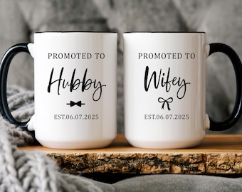 May include: Two white ceramic mugs with black rims and handles. The mugs have black text that reads "Promoted to Hubby EST. 06.07.2025" and "Promoted to Wife EST. 06.07.2025".