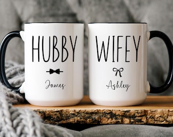 May include: Two white ceramic mugs with black handles and black lettering. The mug on the left says "Hubby James" with a bow tie icon. The mug on the right says "Wifey Ashley" with a bow icon.