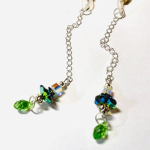 May include: Eyeglass chains with clear loops and silver chains. Each chain has a cluster of iridescent crystals and green beads. The eyeglass chains are designed to be attached to the arms of eyeglasses.