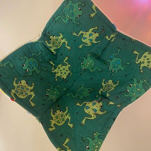 May include: A square, green fabric bowl with a repeating pattern of cartoon frogs in various shades of green. The bowl has a white interior and is likely handmade. The frogs have different patterns and expressions.