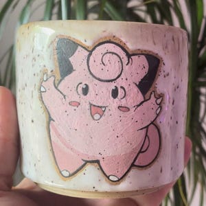 Könnte beinhalten: Keramikbecher mit handgemaltem Design eines rosa Cartoon-Charakters. Der Becher hat einen gesprenkelten, cremefarbenen Hintergrund und einen hellbraunen Rand. Der Charakter hat schwarze Akzente und lächelt mit ausgestreckten Armen.