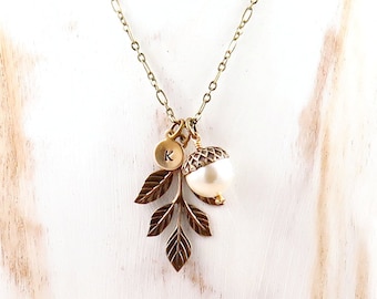 May include: A gold chain necklace with a charm featuring a small acorn, a pearl, and three leaves. The charm also has a small disc with the letter 'K' on it.
