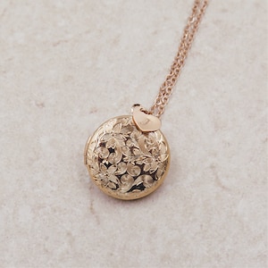May include: Gold locket necklace with a floral design and a small heart charm with the letter 'J' engraved on it.