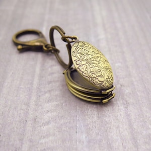 May include: An oval-shaped antique brass locket keychain featuring an intricate floral engraving on the front cover. The locket is attached to a circular split ring and a lobster claw clasp, both finished in a matching antique brass tone.