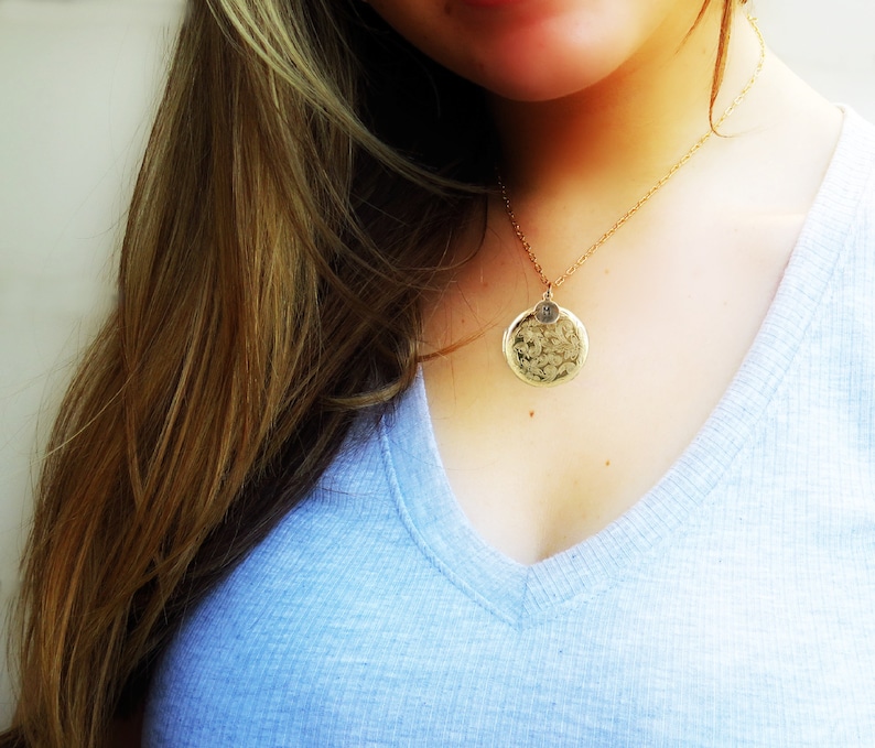 May include: A gold necklace with a round pendant featuring a textured design. The pendant has a small letter charm attached to it.