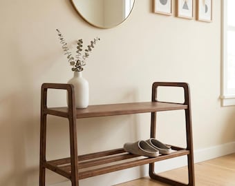 Può includere: Una scarpiera in legno scuro a due livelli dal design minimalista. Un paio di pantofole grigie poggiano sul ripiano inferiore. Un vaso bianco con della vegetazione è posto sul ripiano superiore. Uno specchio rotondo dorato e opere d'arte incorniciate sono appesi al muro.