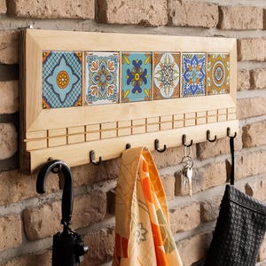 Könnte beinhalten: Ein hölzerner Wand-Schlüssel- und Mantelhaken mit sechs dekorativen Keramikfliesen. Die Fliesen weisen farbenfrohe Blumenmuster in Blau, Orange und Gelb auf. Unter den Fliesen befinden sich sechs schwarze Metallhaken. Ein Regenschirm, ein Handtuch, Schlüssel und eine Tasche hängen.
