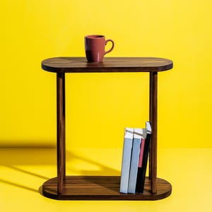 May include: A dark brown wooden side table with an oval top and base, and vertical supports. A maroon mug sits on the table's surface. Three books are stored on the lower shelf. The background is a solid yellow.