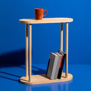 May include: A light wood side table with an oval top and base, supported by four cylindrical legs. An orange mug sits on the table's surface. Three books are placed on the lower shelf. The table is set against a blue background.