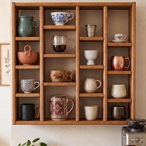 Pu&ograve; includere: Una mensola in legno con una variet&agrave; di tazze e ciotole. La collezione comprende pezzi in ceramica, vetro e metallo in una gamma di colori e motivi. Una piccola pianta e una macchina per l'espresso sono visibili sotto.
