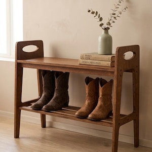 Peut inclure: Un meuble à chaussures en bois à deux étagères. L'étagère supérieure contient un petit vase avec de la verdure et deux livres. L'étagère inférieure présente quatre paires de bottes de cowboy marron. Le meuble a un design simple et rustique.