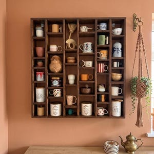 Pu&ograve; includere: Una mensola espositiva in legno marrone scuro piena di una variet&agrave; di tazze e piccoli oggetti decorativi. La mensola &egrave; montata su una parete color pesca. Sono visibili anche un portapiante in macram&egrave; e una piccola teiera in ottone.