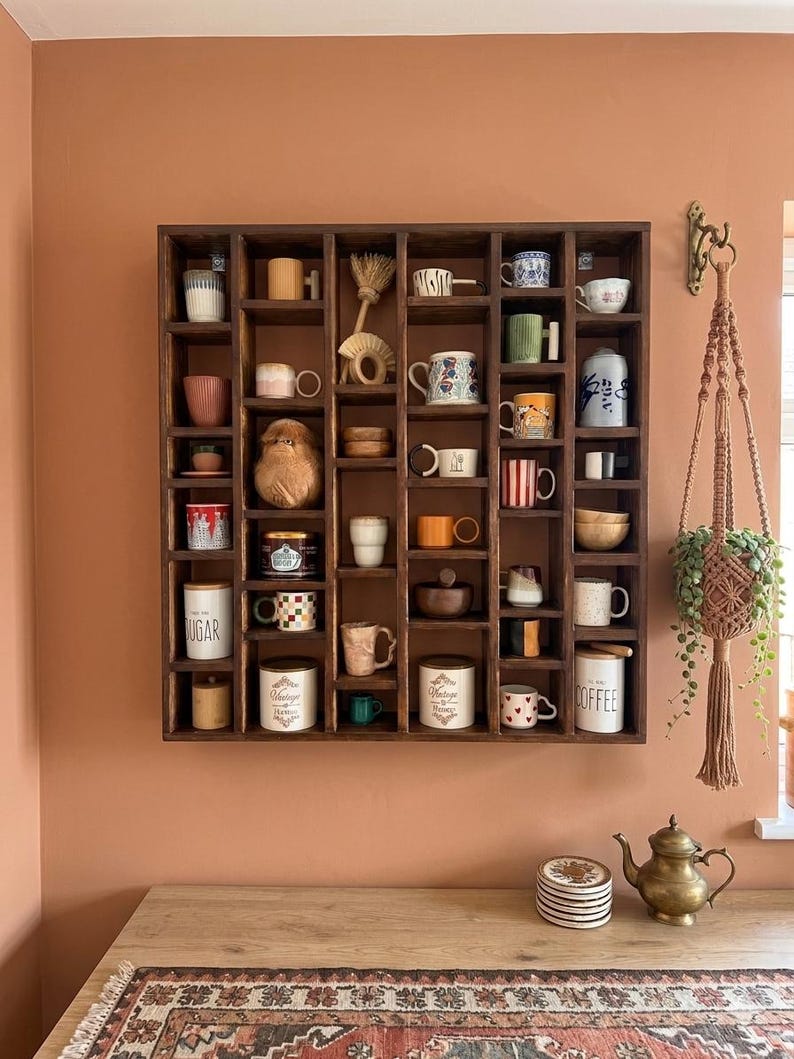 Pu&ograve; includere: Una mensola espositiva in legno marrone scuro piena di una variet&agrave; di tazze e piccoli oggetti decorativi. La mensola &egrave; montata su una parete color pesca. Sono visibili anche un portapiante in macram&egrave; e una piccola teiera in ottone.