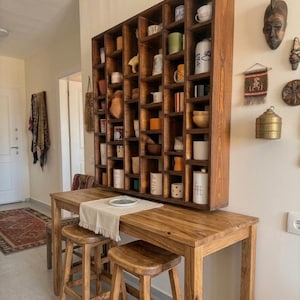 Pu&ograve; includere: Un set da cucina in legno con una grande mensola scura piena di tazze e oggetti decorativi. Sotto, un tavolo in legno abbinato con due sgabelli &egrave; apparecchiato con un runner neutro e un piatto. L'ambiente &egrave; caldo e accogliente.