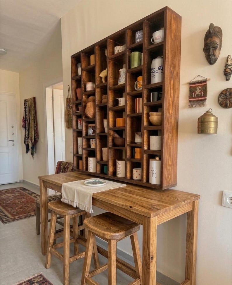 Pu&ograve; includere: Un set da cucina in legno con una grande mensola scura piena di tazze e oggetti decorativi. Sotto, un tavolo in legno abbinato con due sgabelli &egrave; apparecchiato con un runner neutro e un piatto. L'ambiente &egrave; caldo e accogliente.